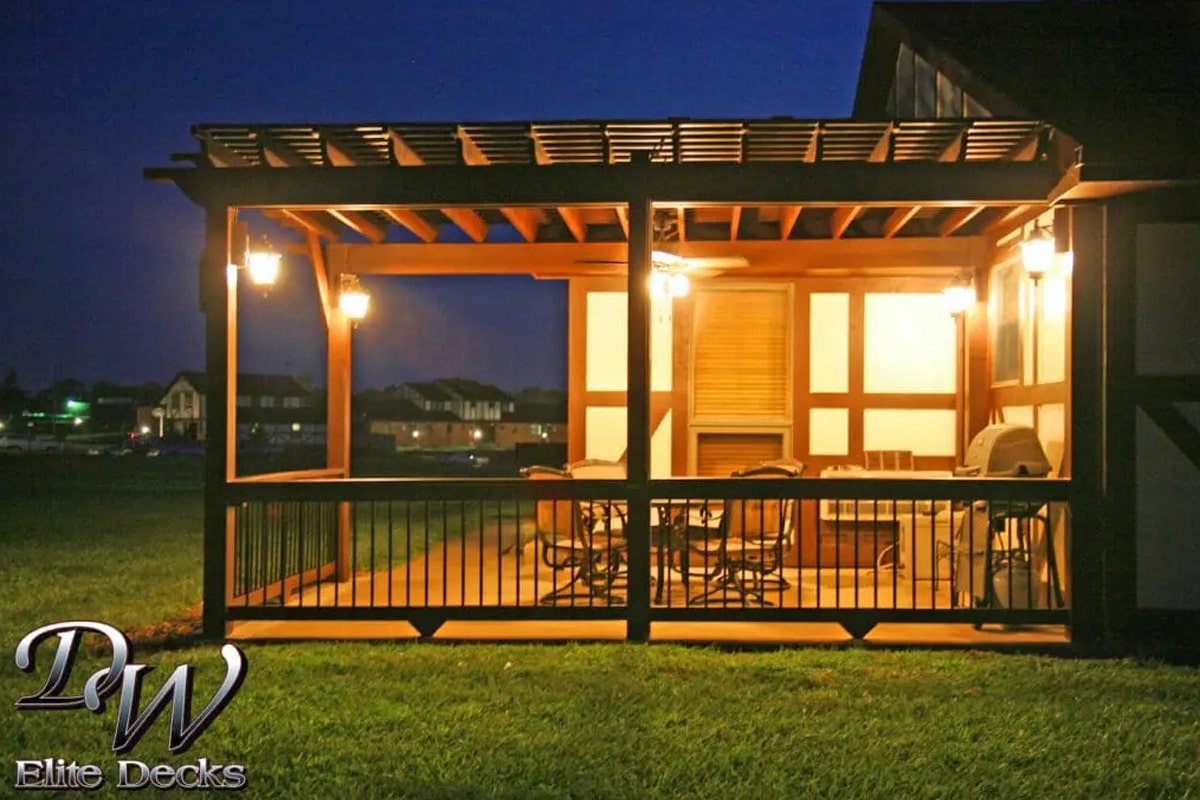 Covered deck with a pergola roof and lit lantern fixtures at night, with patio furniture inside and a grill on the right